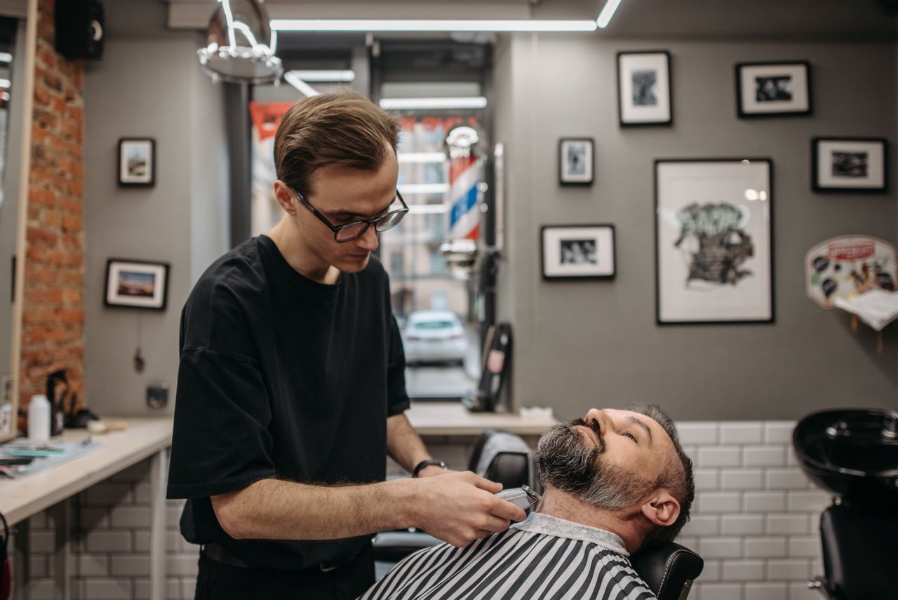 our-mission A barber meticulously trims a bearded man's hair in a contemporary barbershop setting.